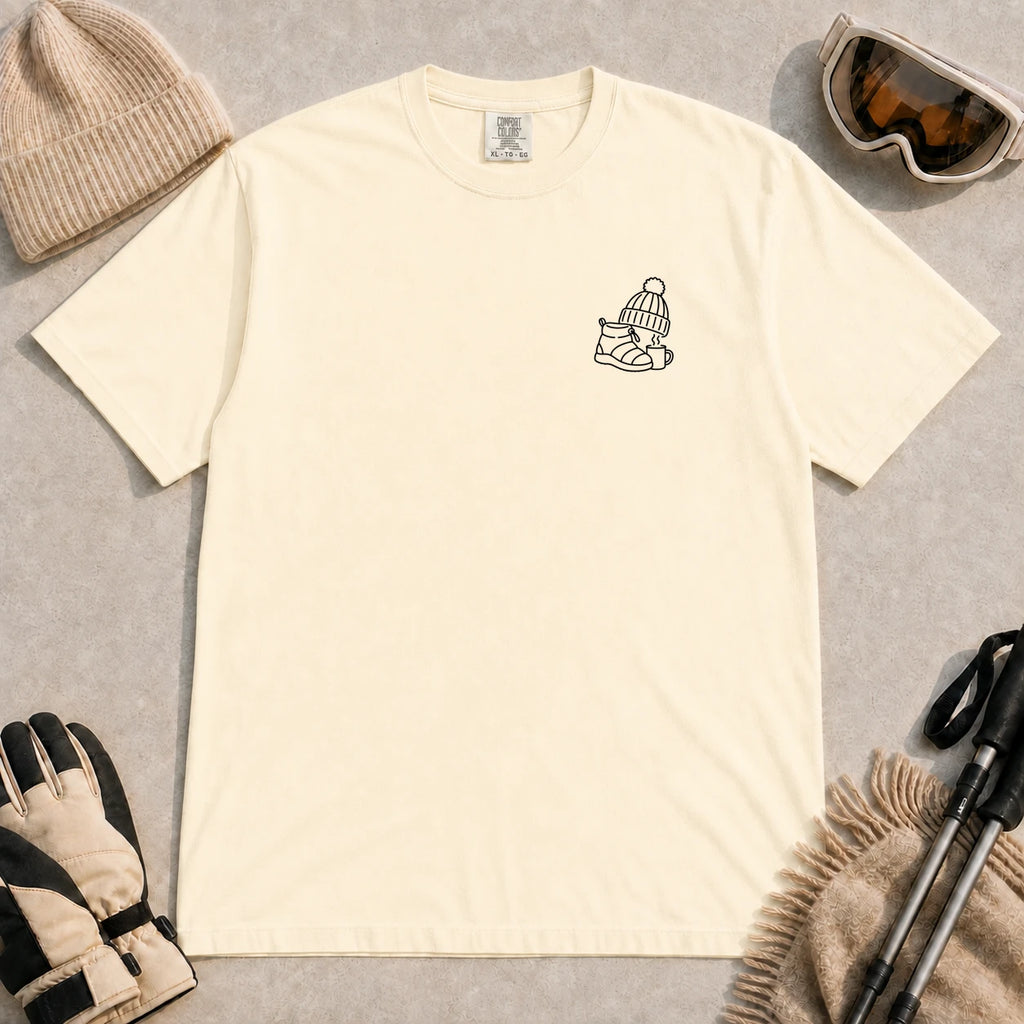 Ivory tee featuring a minimalist black line-art design of a ski boot, winter beanie, and steaming mug on the left chest.