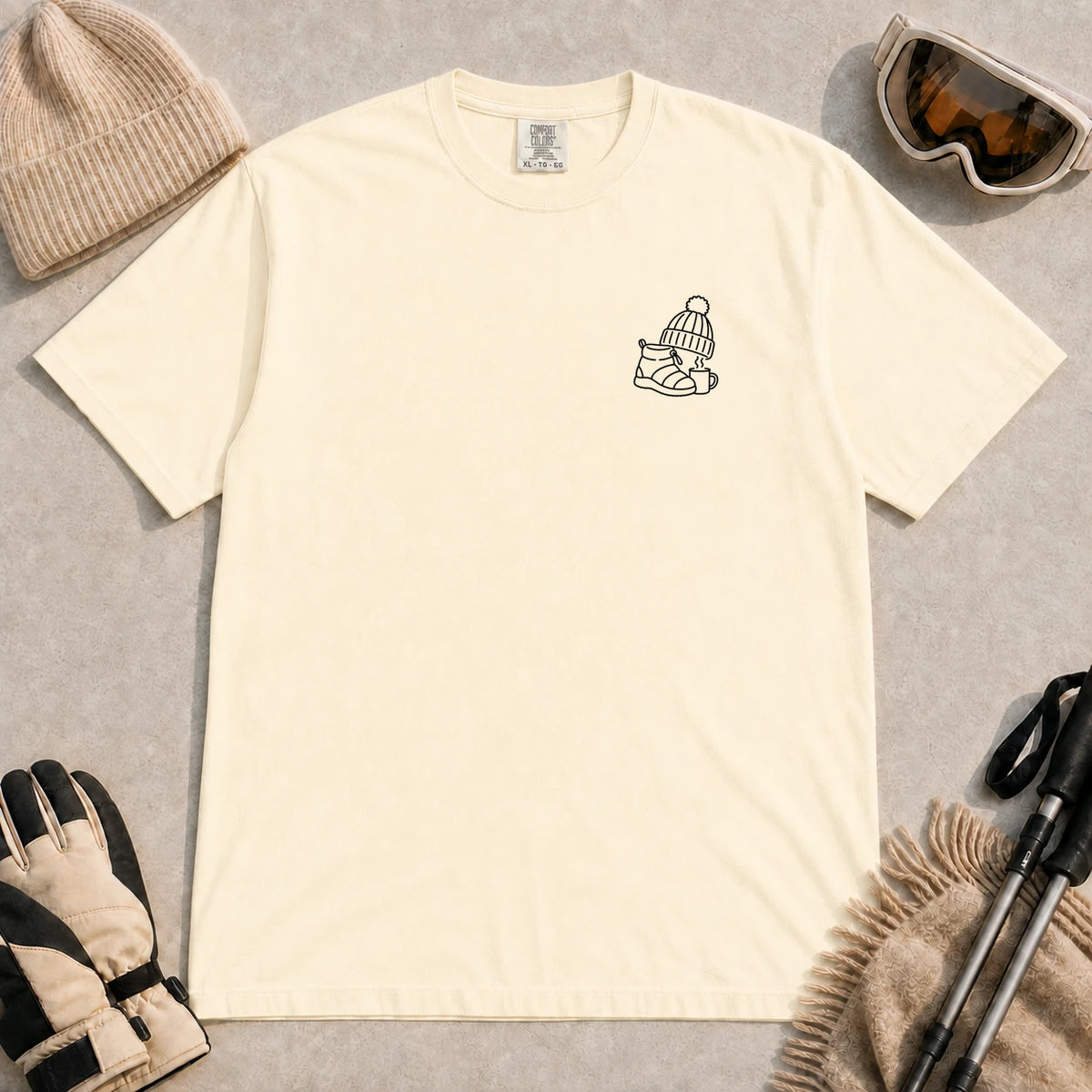 Ivory tee featuring a minimalist black line-art design of a ski boot, winter beanie, and steaming mug on the left chest.