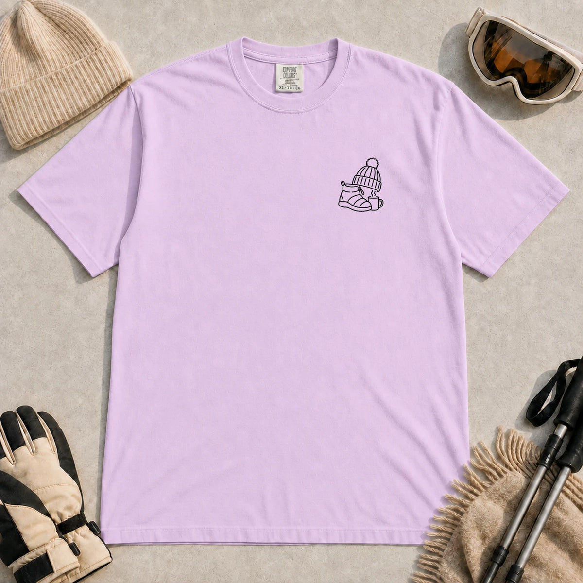 Orchid tee featuring a minimalist black line-art design of a ski boot, winter beanie, and steaming mug on the left chest.