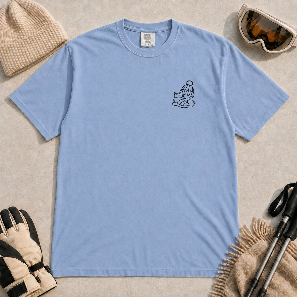 Washed Denim tee featuring a minimalist black line-art design of a ski boot, winter beanie, and steaming mug on the left chest.