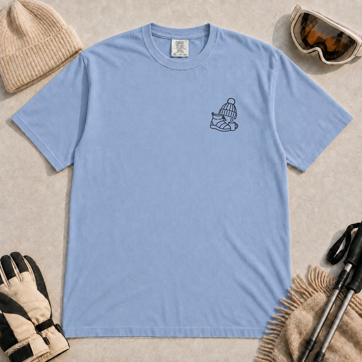 Washed Denim tee featuring a minimalist black line-art design of a ski boot, winter beanie, and steaming mug on the left chest.