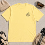 Cornsilk tee featuring a minimalist black line-art design of a ski boot, winter beanie, and steaming mug on the left chest.
