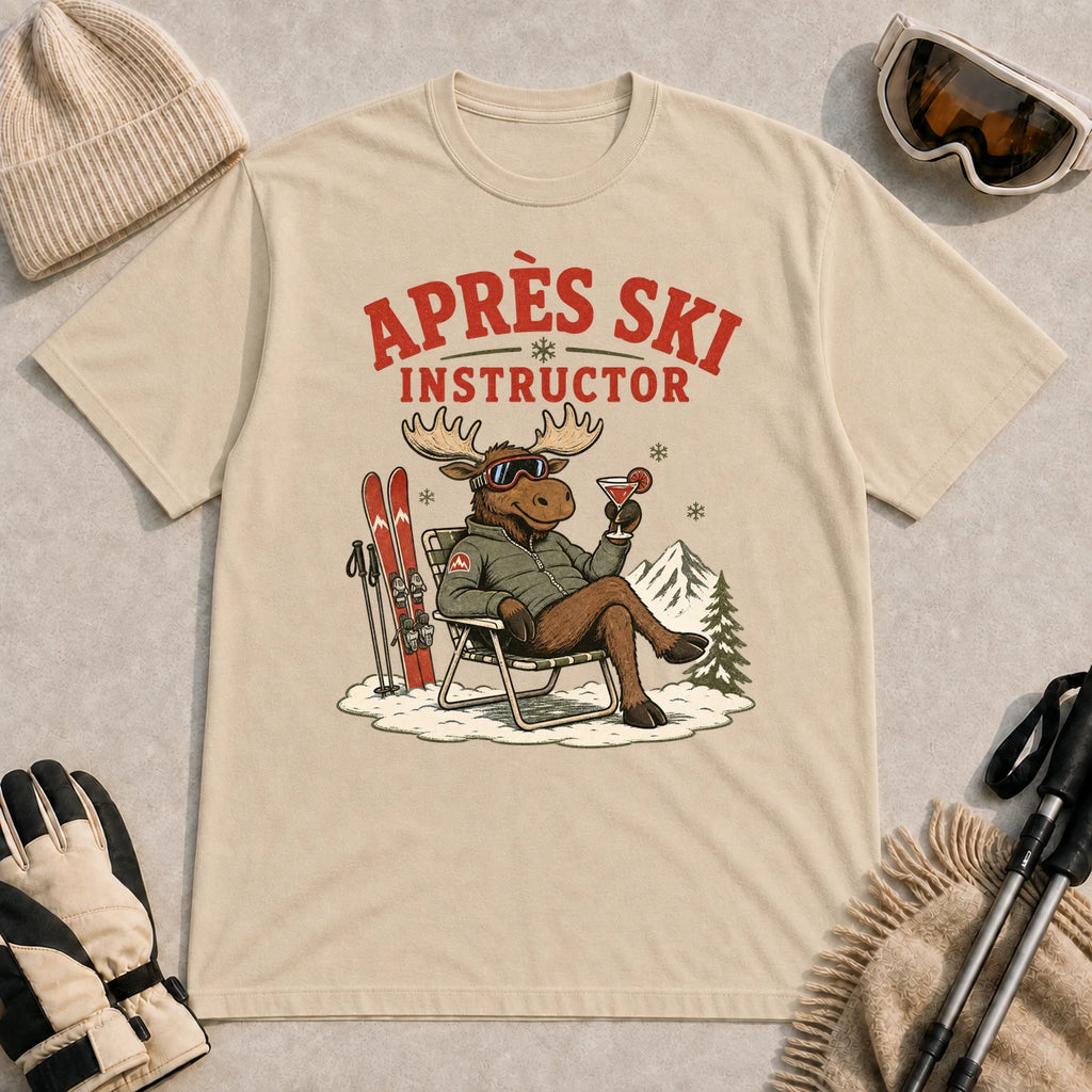 Sand tee featuring a relaxed moose lounging in a chair beside skis and holding a cocktail, with the phrase “Après Ski Instructor.”