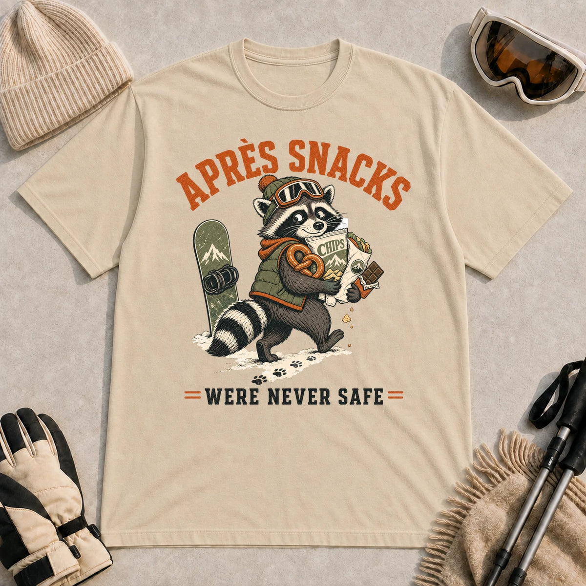 Sand tee featuring a snack-loaded raccoon in ski goggles carrying chips, pretzels, and chocolate beside a snowboard, with the phrase “Après Snacks Were Never Safe.”