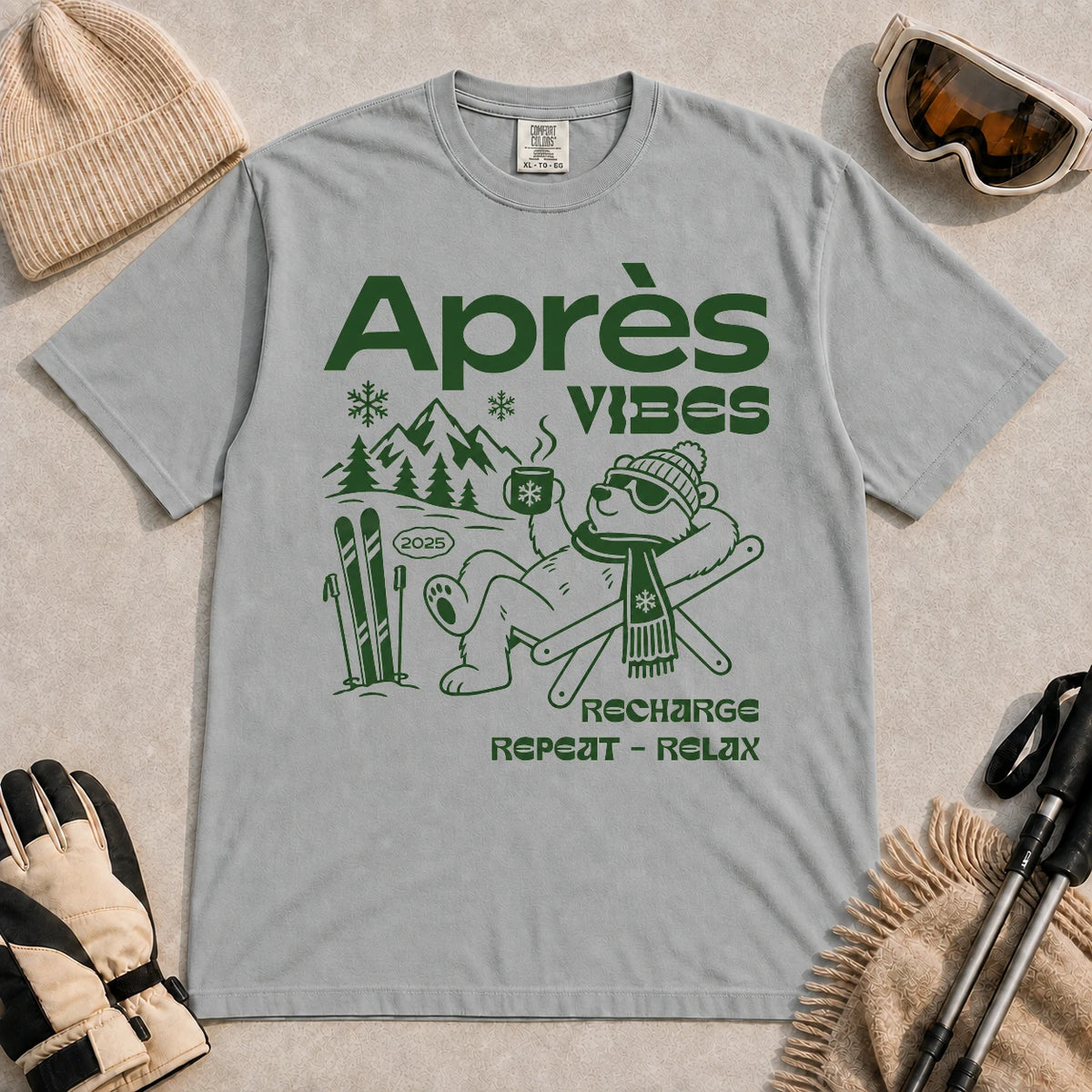 Granite tee featuring a relaxed bear in sunglasses holding a steaming mug beside skis, with retro green “Après Vibes” and “Recharge, Repeat – Relax” text.