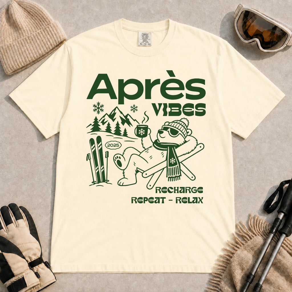 Ivory tee featuring a relaxed bear in sunglasses holding a steaming mug beside skis, with retro green “Après Vibes” and “Recharge, Repeat – Relax” text.