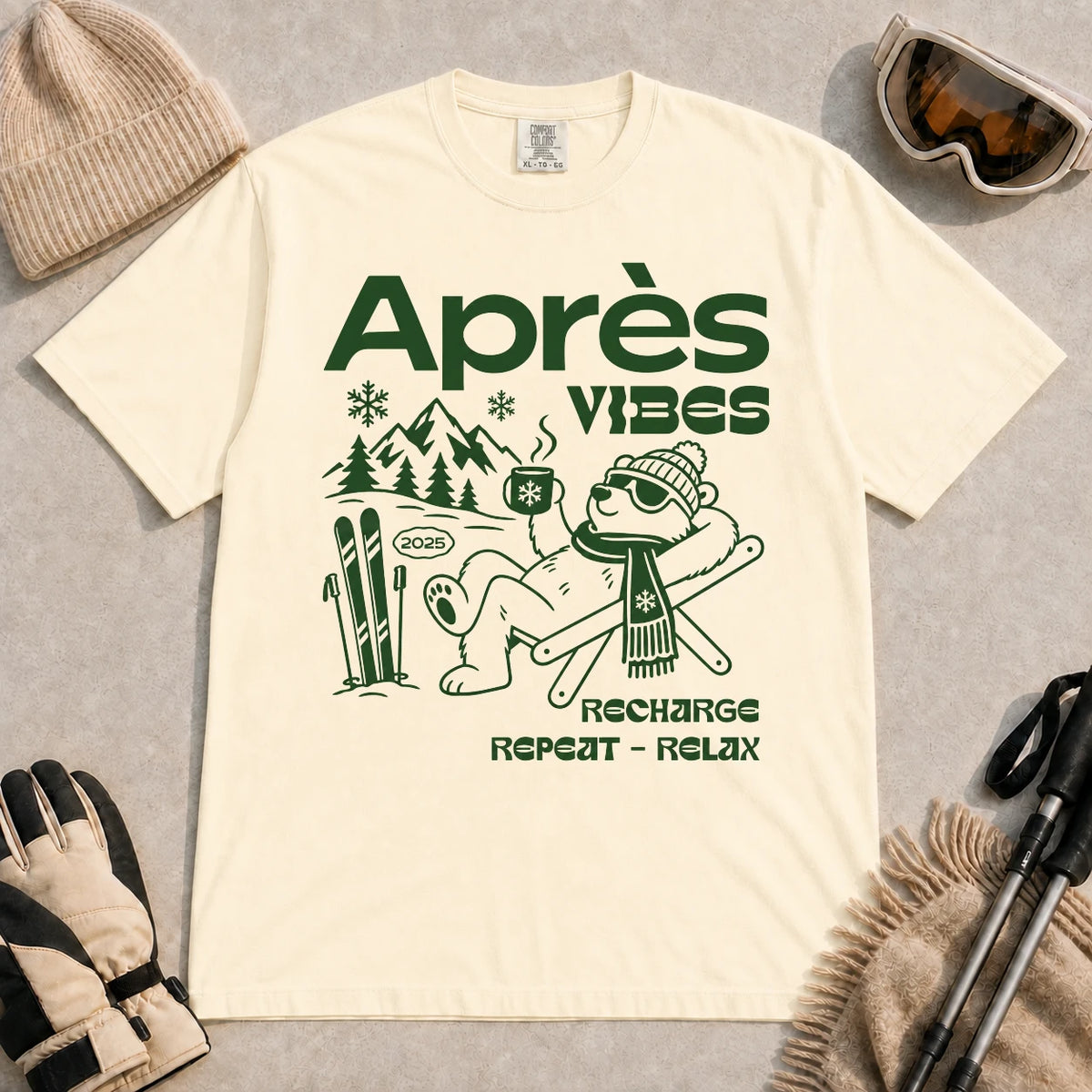 Ivory tee featuring a relaxed bear in sunglasses holding a steaming mug beside skis, with retro green “Après Vibes” and “Recharge, Repeat – Relax” text.