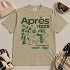 Khaki tee featuring a relaxed bear in sunglasses holding a steaming mug beside skis, with retro green “Après Vibes” and “Recharge, Repeat – Relax” text.