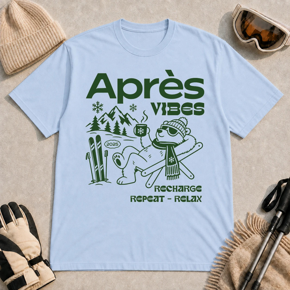 Light blue tee featuring a relaxed bear in sunglasses holding a steaming mug beside skis, with retro green “Après Vibes” and “Recharge, Repeat – Relax” text."