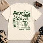 Natural tee featuring a relaxed bear in sunglasses holding a steaming mug beside skis, with retro green “Après Vibes” and “Recharge, Repeat – Relax” text.