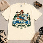 Ivory tee featuring a cheerful bear snowboarding past a blue-square trail sign above the phrase “Blue Square Enthusiast” with “Groomers. Good Vibes. More Runs.”