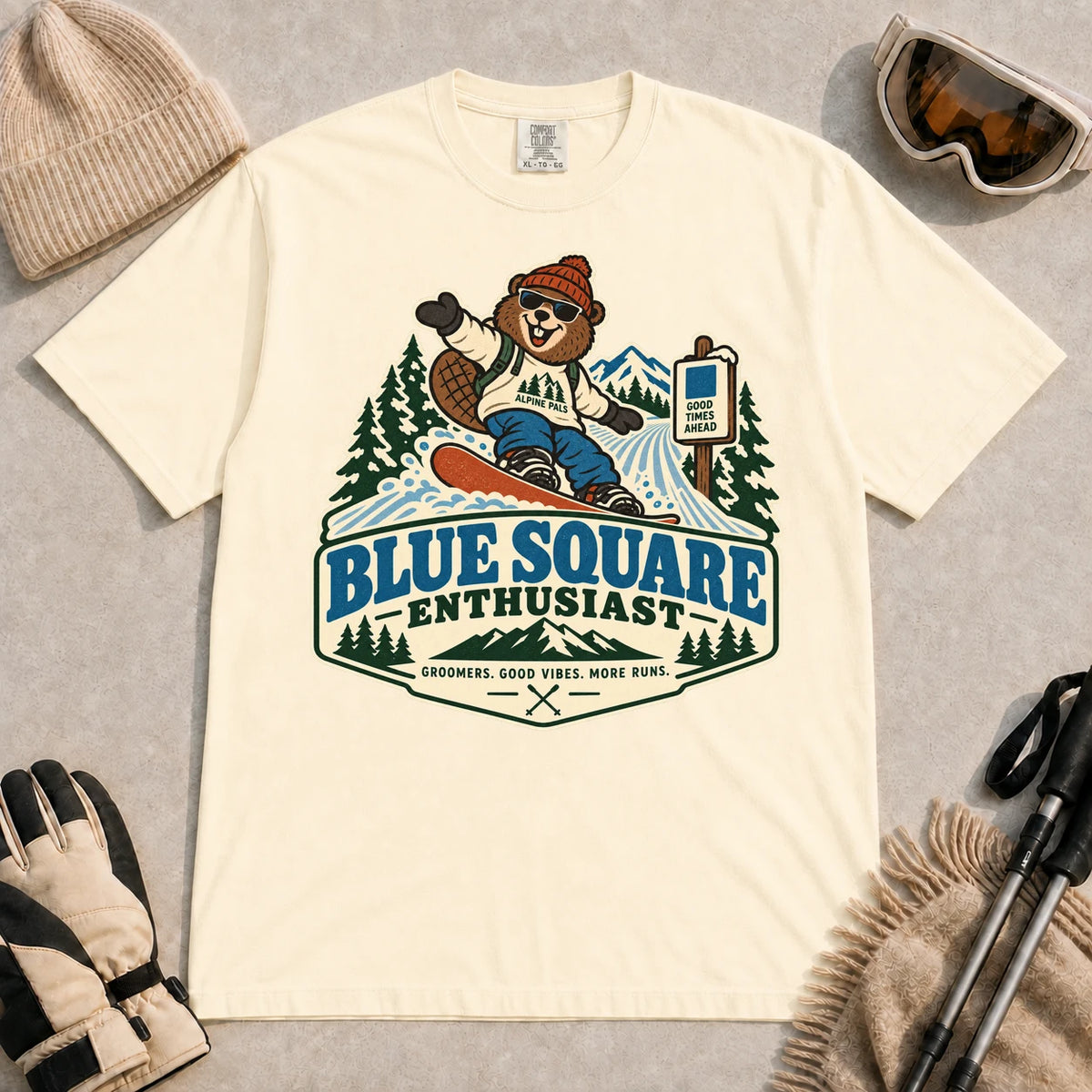 Ivory tee featuring a cheerful bear snowboarding past a blue-square trail sign above the phrase “Blue Square Enthusiast” with “Groomers. Good Vibes. More Runs.”