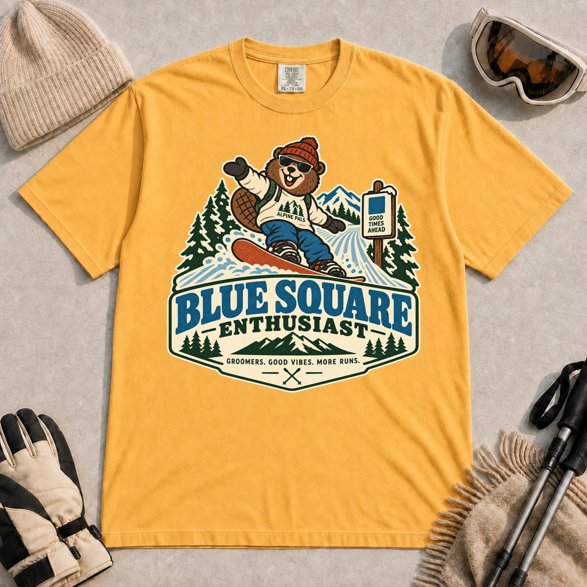 Mustard tee featuring a cheerful bear snowboarding past a blue-square trail sign above the phrase “Blue Square Enthusiast” with “Groomers. Good Vibes. More Runs.”