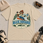 Sand tee featuring a cheerful bear snowboarding past a blue-square trail sign above the phrase “Blue Square Enthusiast” with “Groomers. Good Vibes. More Runs.”