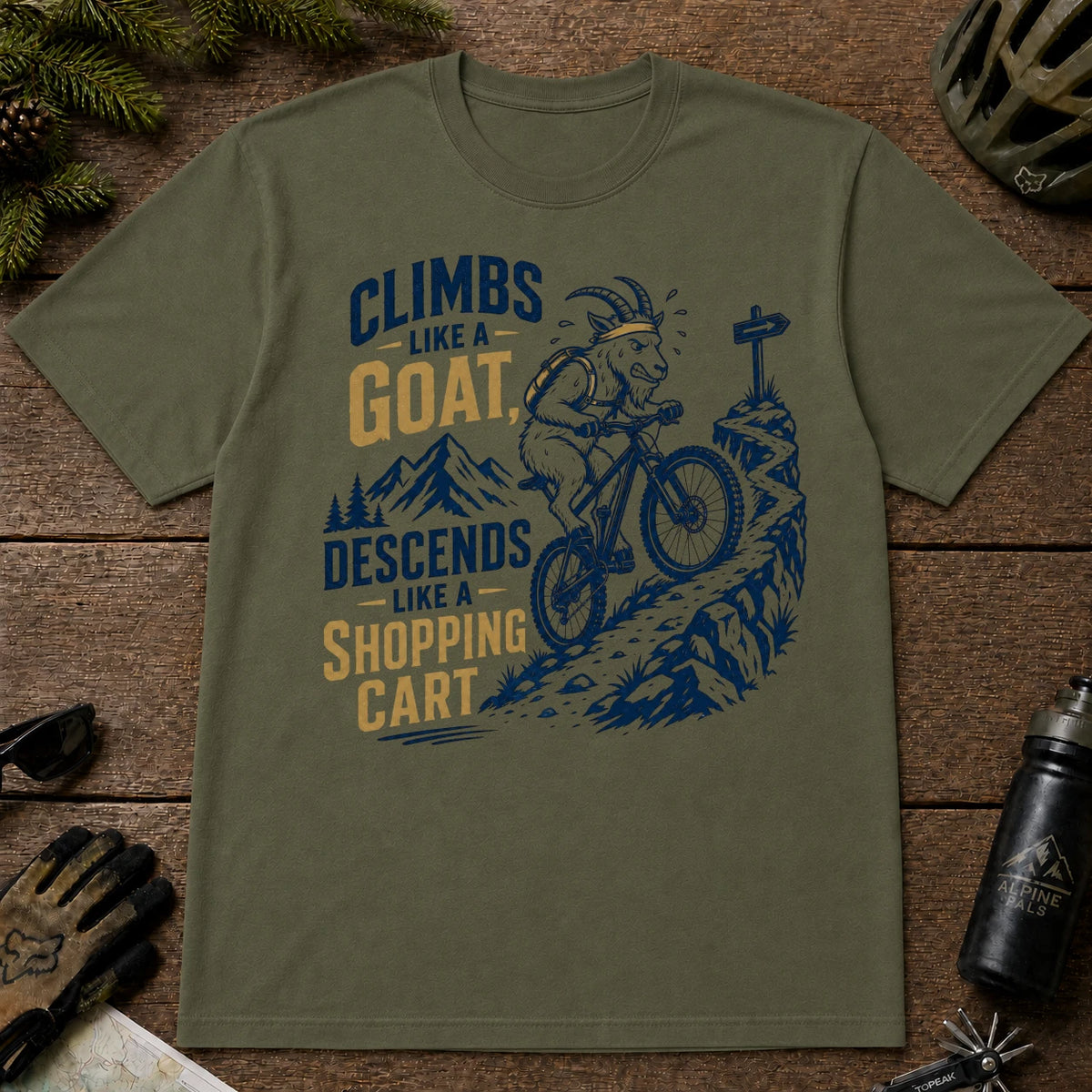 Military Green tee featuring a mountain goat riding a mountain bike down a rocky trail, with the phrase “Climbs Like a Goat, Descends Like a Shopping Cart.”