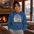 Woman wearing a blue sweatshirt with a mountain and dog design, standing in a cozy room with a fireplace and snowy window.