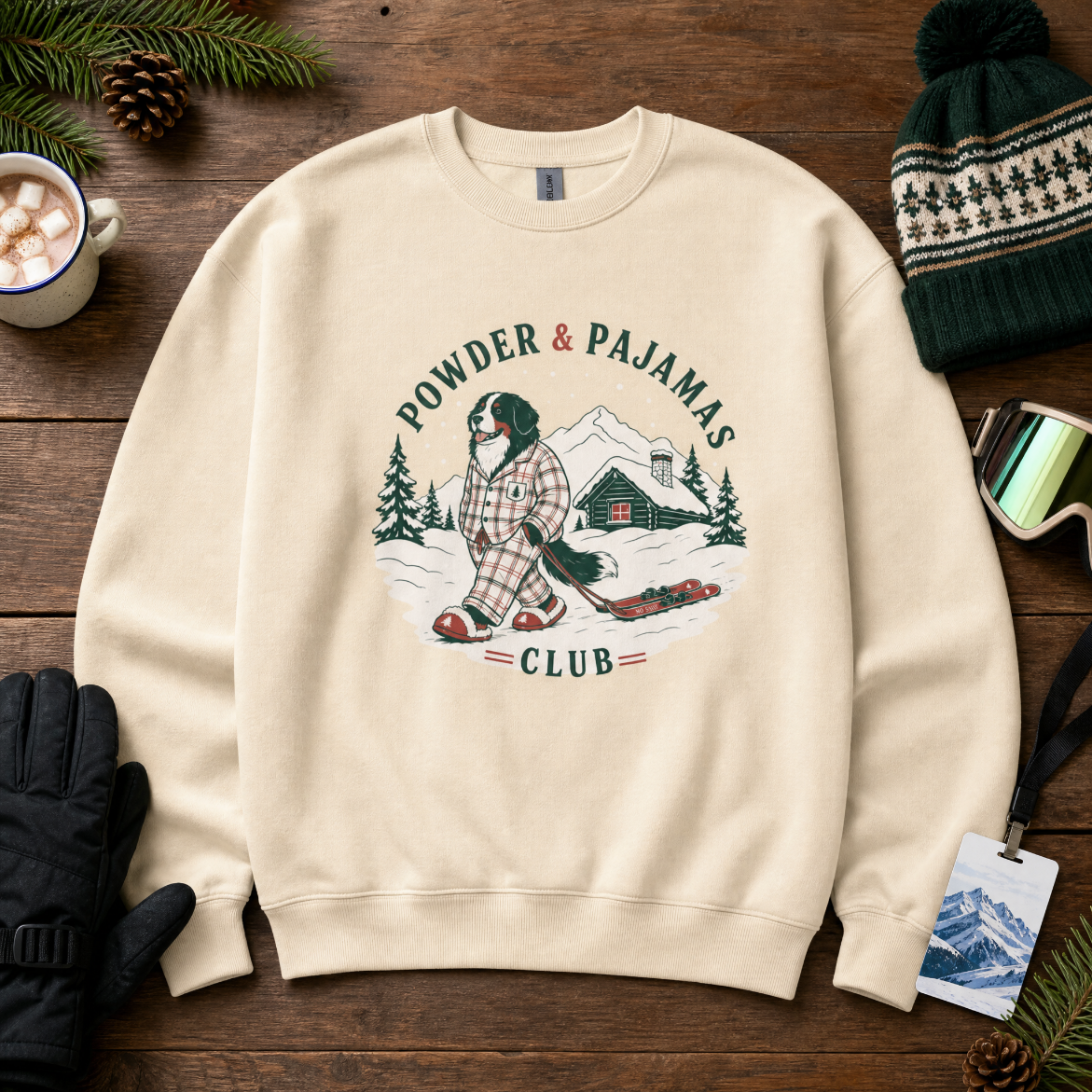 Sand colored sweatshirt with 'Powder & Pajamas Club' design on a wooden surface with winter accessories.