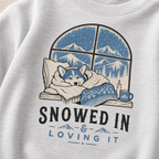 Ash gray sweatshirt with a graphic of a dog by a window with mountains, text 'SNOWED IN & LOVING IT', and a cup of coffee.