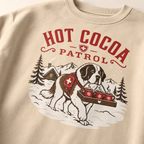 Sand colored sweatshirt with a graphic of a dog carrying hot cocoa mugs and 'Hot Cocoa Patrol' text.