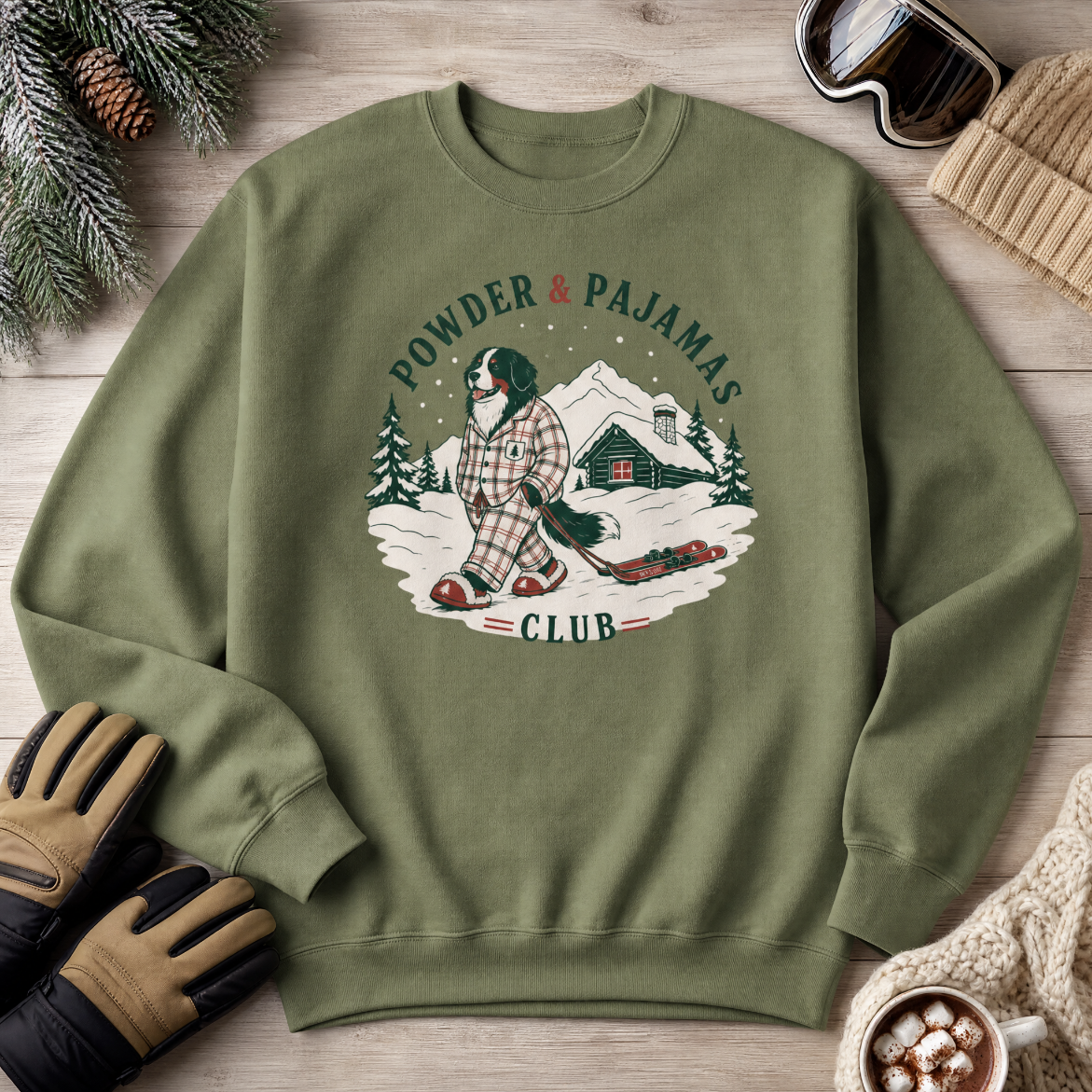 Military green sweatshirt with a winter graphic and text on a wooden surface with winter accessories.