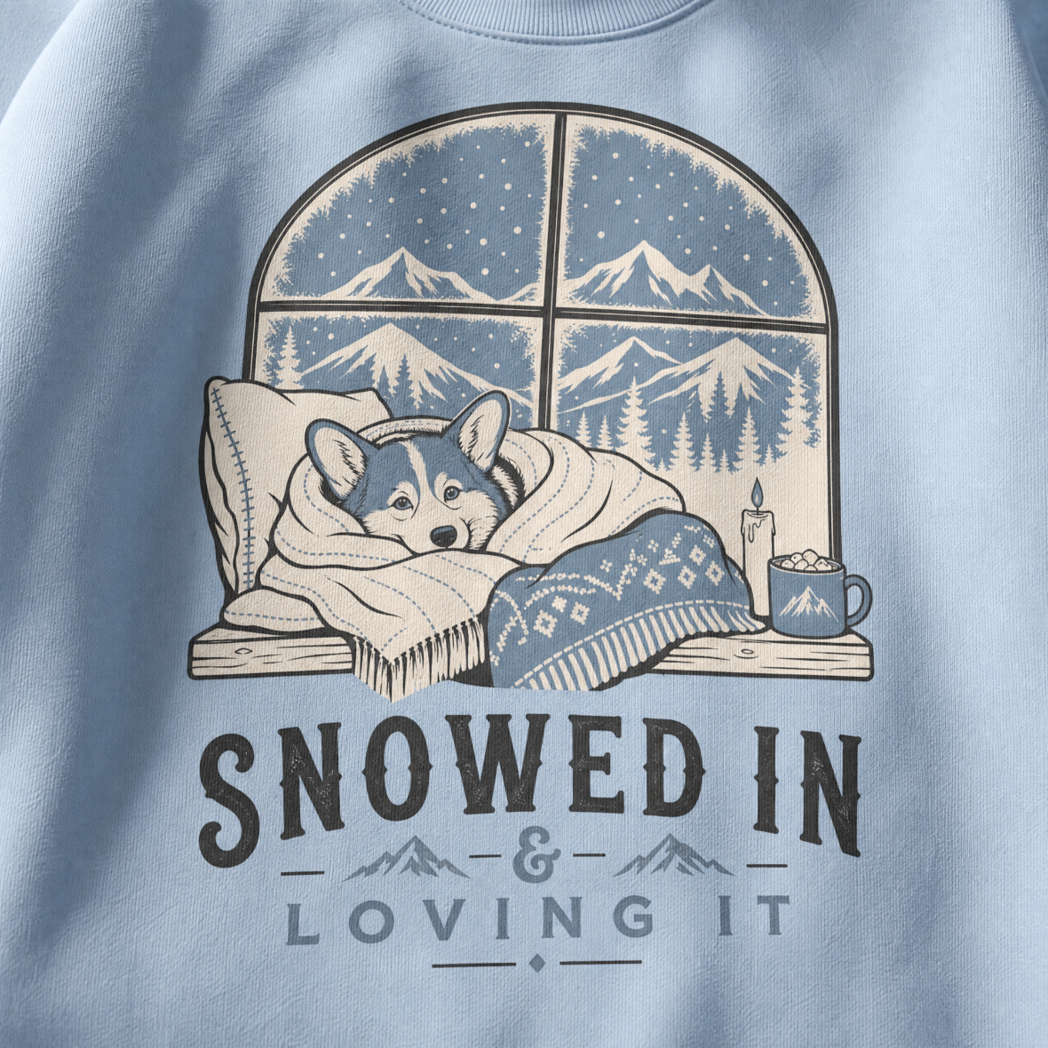 Light blue sweatshirt with a graphic of a dog by a window with mountains, text 'SNOWED IN & LOVING IT'.