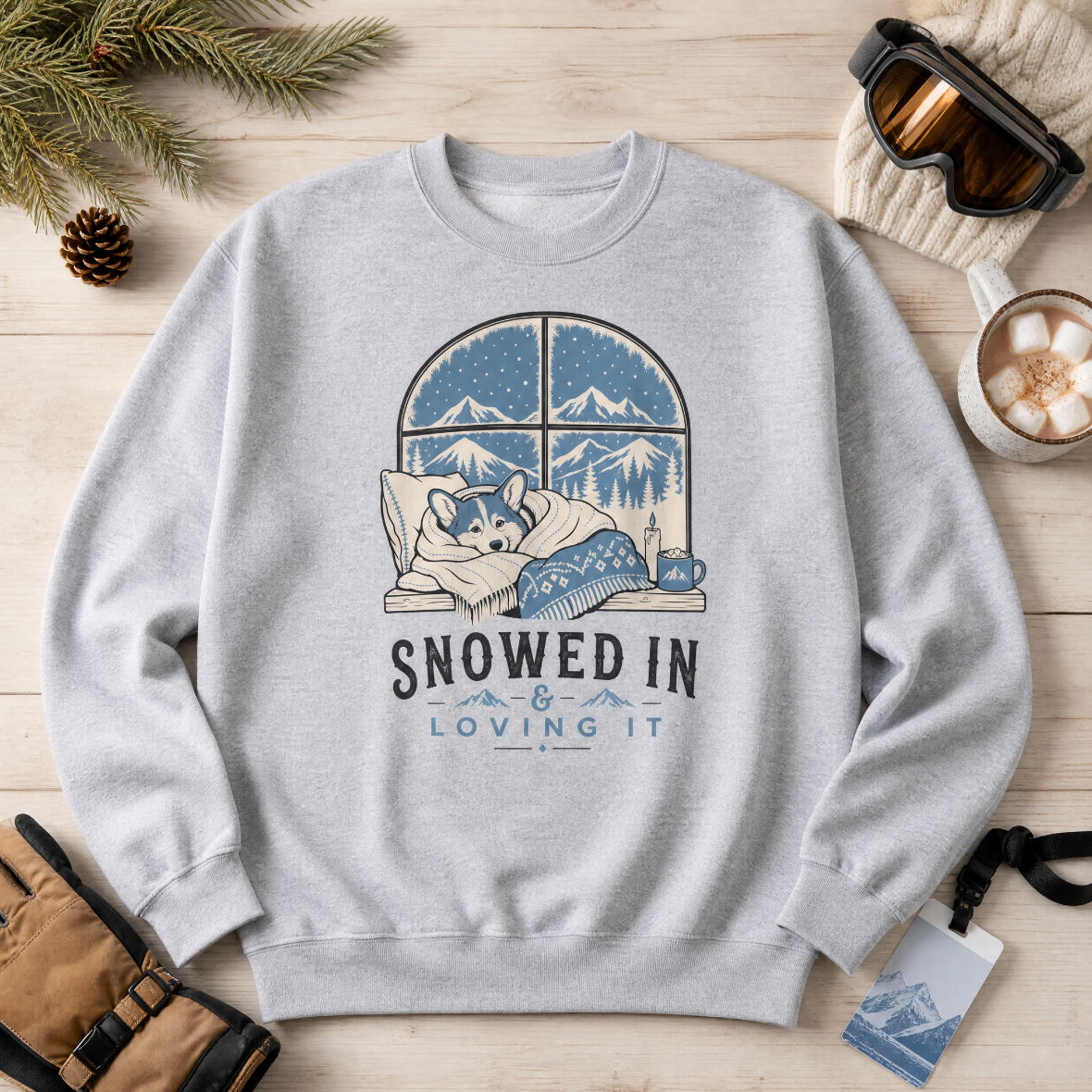Sport gray sweatshirt with a winter-themed design featuring a dog and text 'Snowed In & Loving It' on a wooden surface with winter accessories.