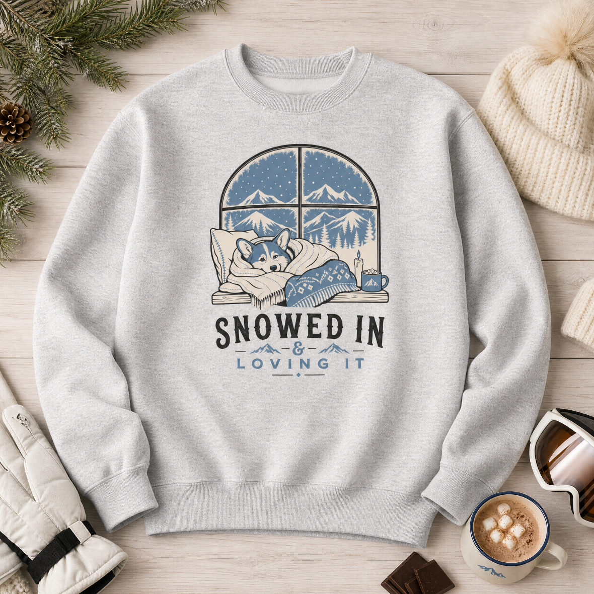 Ash gray sweatshirt with a graphic of a dog by a window and 'Snowed In & Loving It' text, placed on a wooden surface with winter accessories.