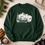 Forest green sweatshirt with a mountain graphic on a textured surface with a cup of coffee and pine branches.