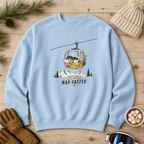 Light blue sweatshirt with a ski lift graphic and text on a wooden surface with winter accessories.