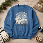 Indigo blue sweatshirt with a graphic of a dog by a window and 'Snowed In & Loving It' text, surrounded by winter accessories on a wooden surface.