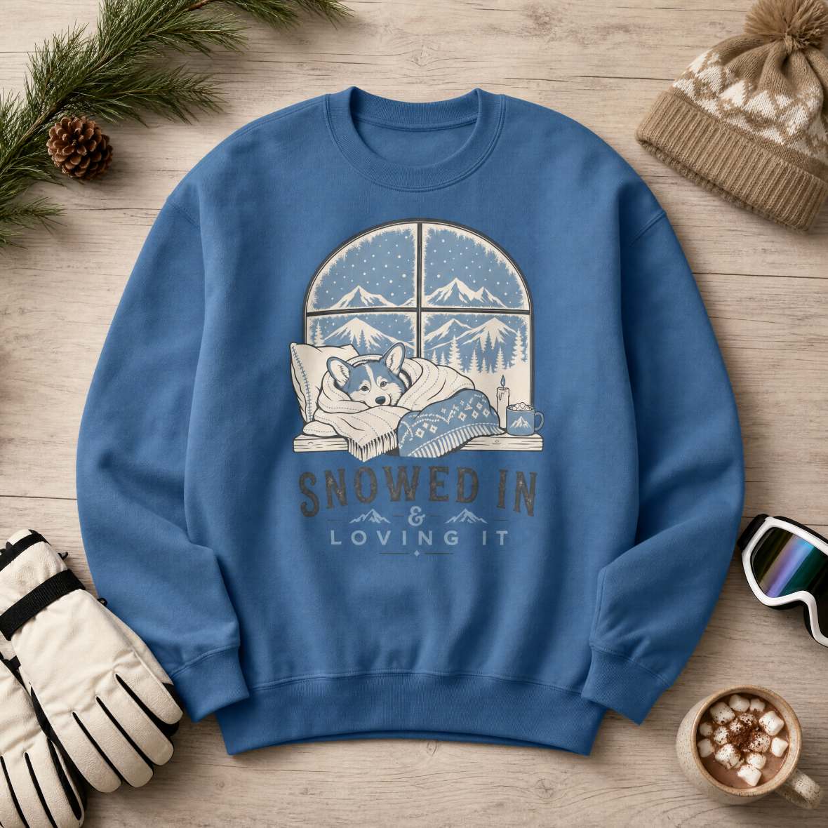 Indigo blue sweatshirt with a graphic of a dog by a window and 'Snowed In & Loving It' text, surrounded by winter accessories on a wooden surface.