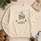 Sand colored sweatshirt with a graphic design of a ski lift and text, surrounded by winter accessories on a wooden surface.