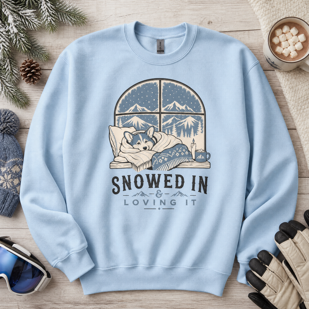 Light blue sweatshirt with a winter-themed design featuring a dog and text 'Snowed In & Loving It' on a wooden surface with winter accessories.