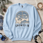 Light blue sweatshirt with a winter-themed design featuring a dog and text 'Snowed In & Loving It' on a wooden surface with winter accessories.
