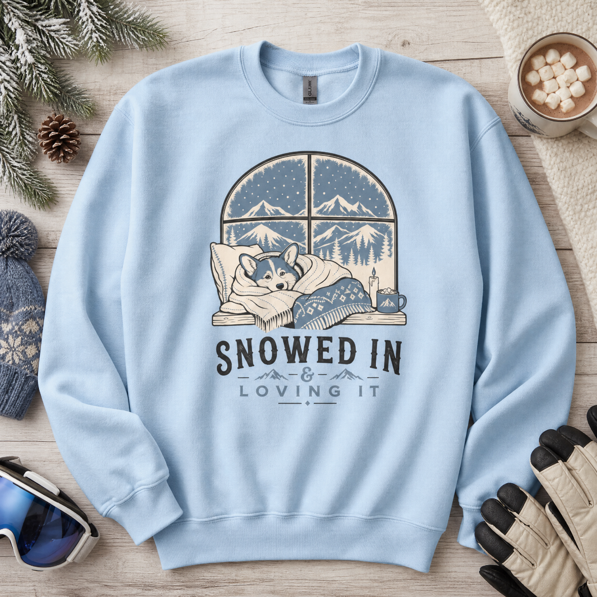 Light blue sweatshirt with a winter-themed design featuring a dog and text 'Snowed In & Loving It' on a wooden surface with winter accessories.