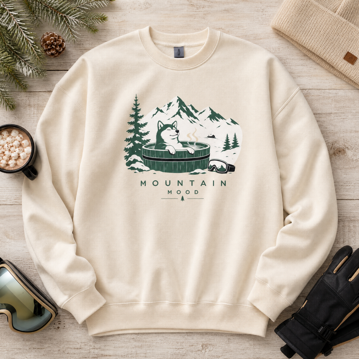 Sand colored sweatshirt with mountain graphic and text on a wooden surface with winter accessories.
