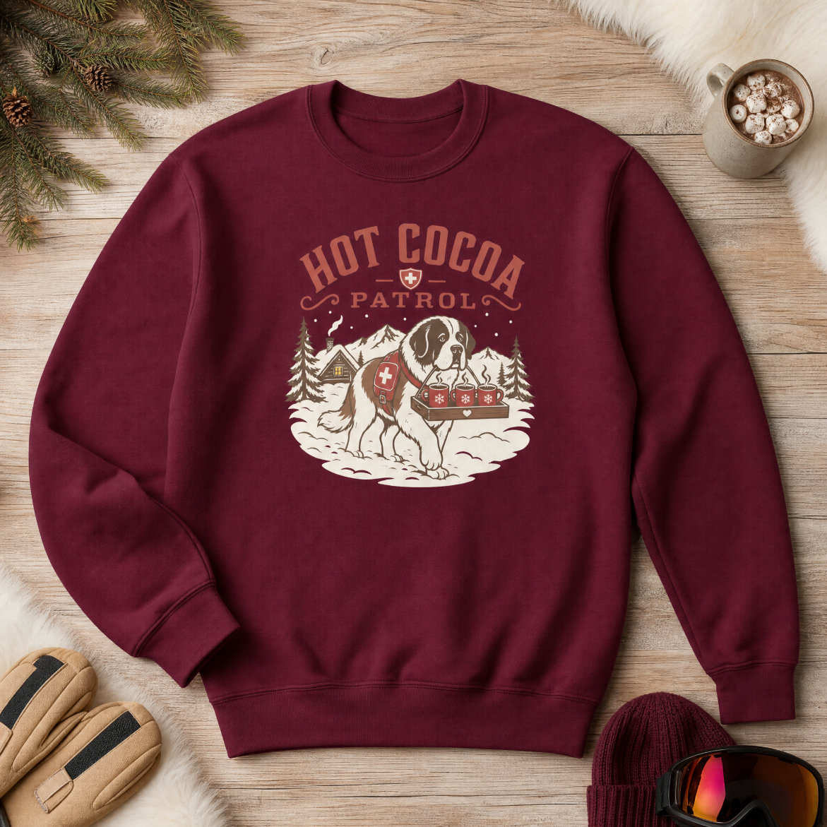 Maroon sweatshirt with 'Hot Cocoa Patrol' graphic on a wooden surface with winter accessories.