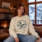 Woman wearing a 'Powder & Pajamas Club' sweatshirt in a cozy living room with a fireplace and snowy mountain view.