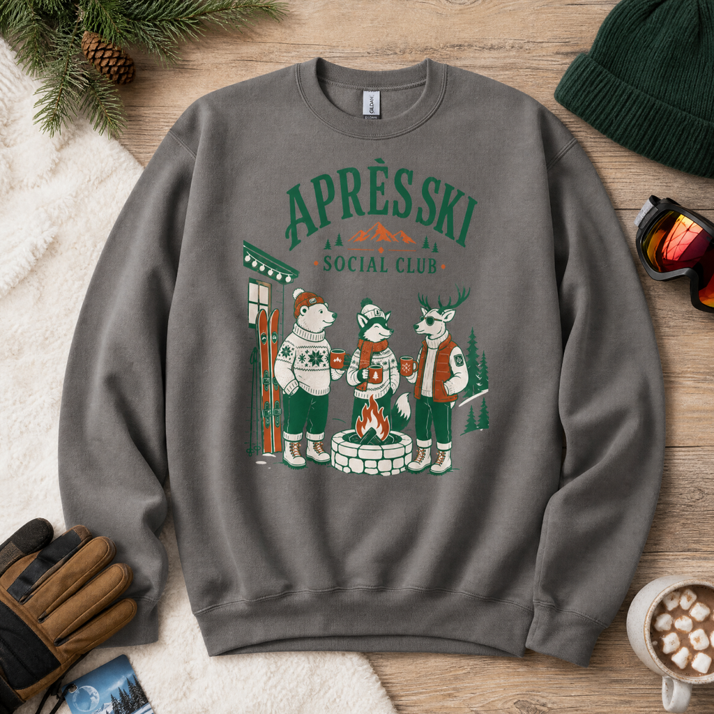 Charcoal gray sweatshirt with 'APRES SKI SOCIAL CLUB' design featuring cartoon characters around a campfire, placed on a wooden surface with winter accessories.