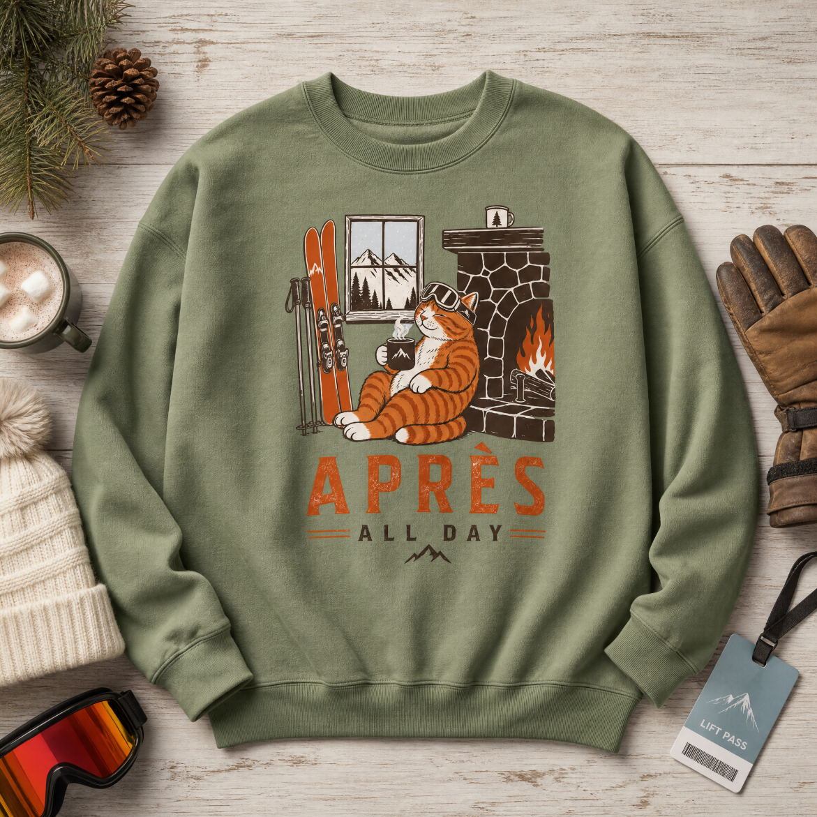Military green sweatshirt with cat design and 'Apres All Day' text on a wooden surface with winter accessories.
