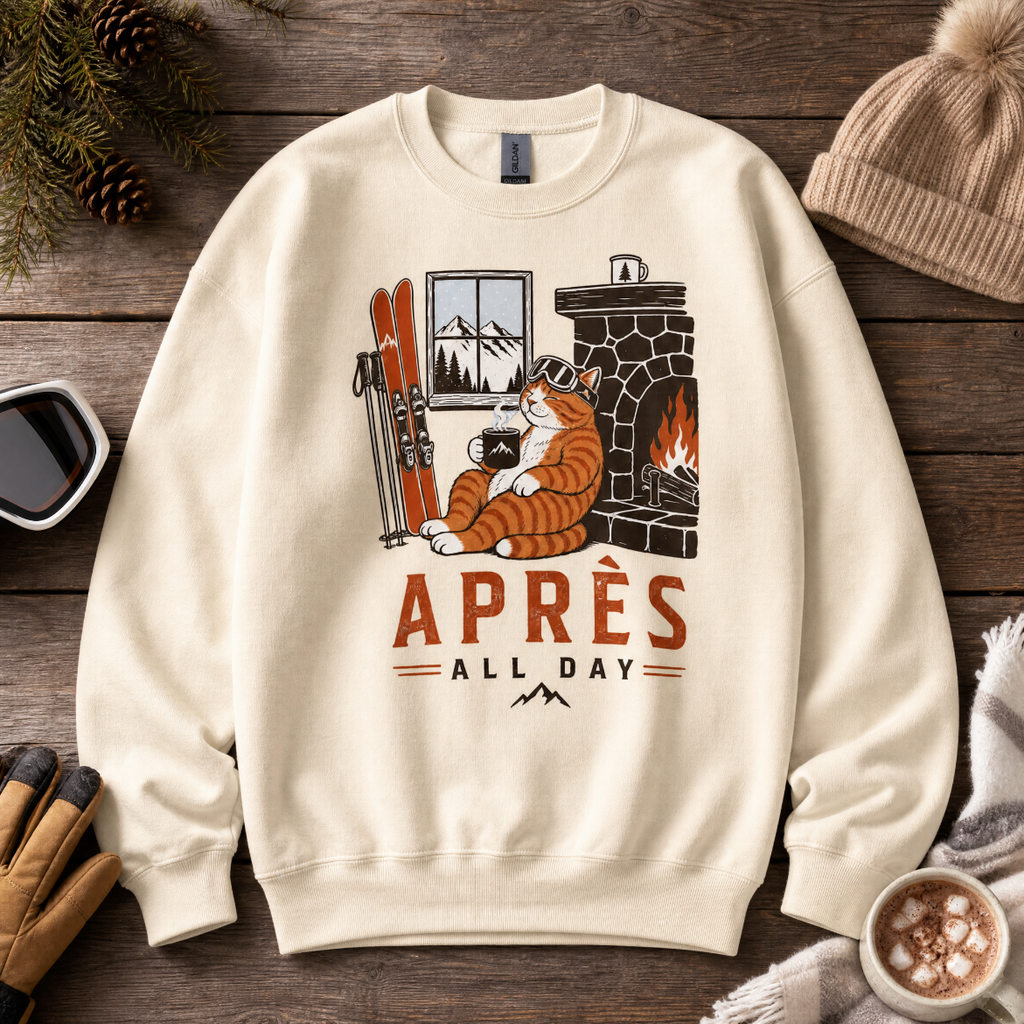 Sand colored sweatshirt with a graphic of a cat by a fireplace and 'APRES ALL DAY' text, placed on a wooden surface with winter accessories.