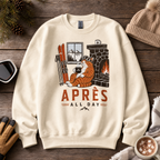 Sand colored sweatshirt with a graphic of a cat by a fireplace and 'APRES ALL DAY' text, placed on a wooden surface with winter accessories.