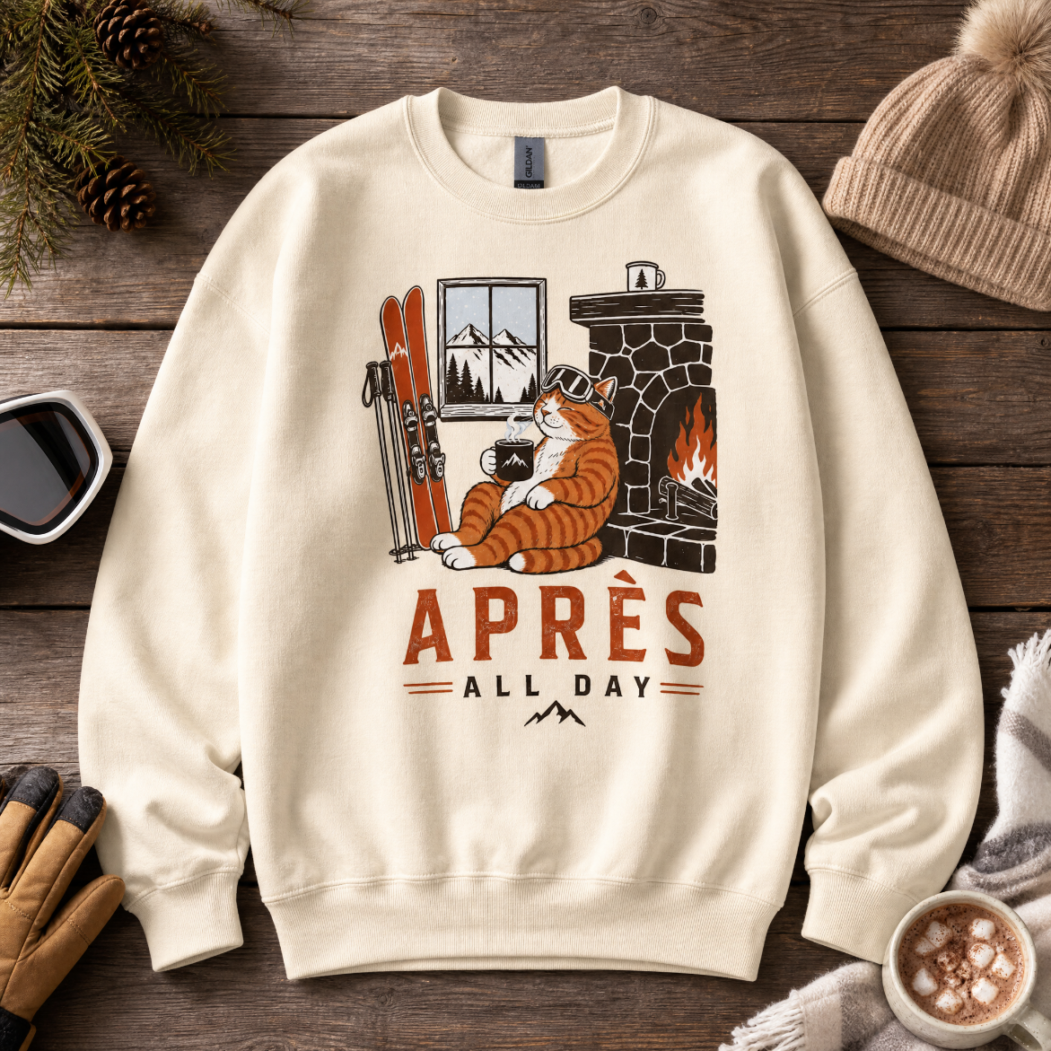 Sand colored sweatshirt with a graphic of a cat by a fireplace and 'APRES ALL DAY' text, placed on a wooden surface with winter accessories.