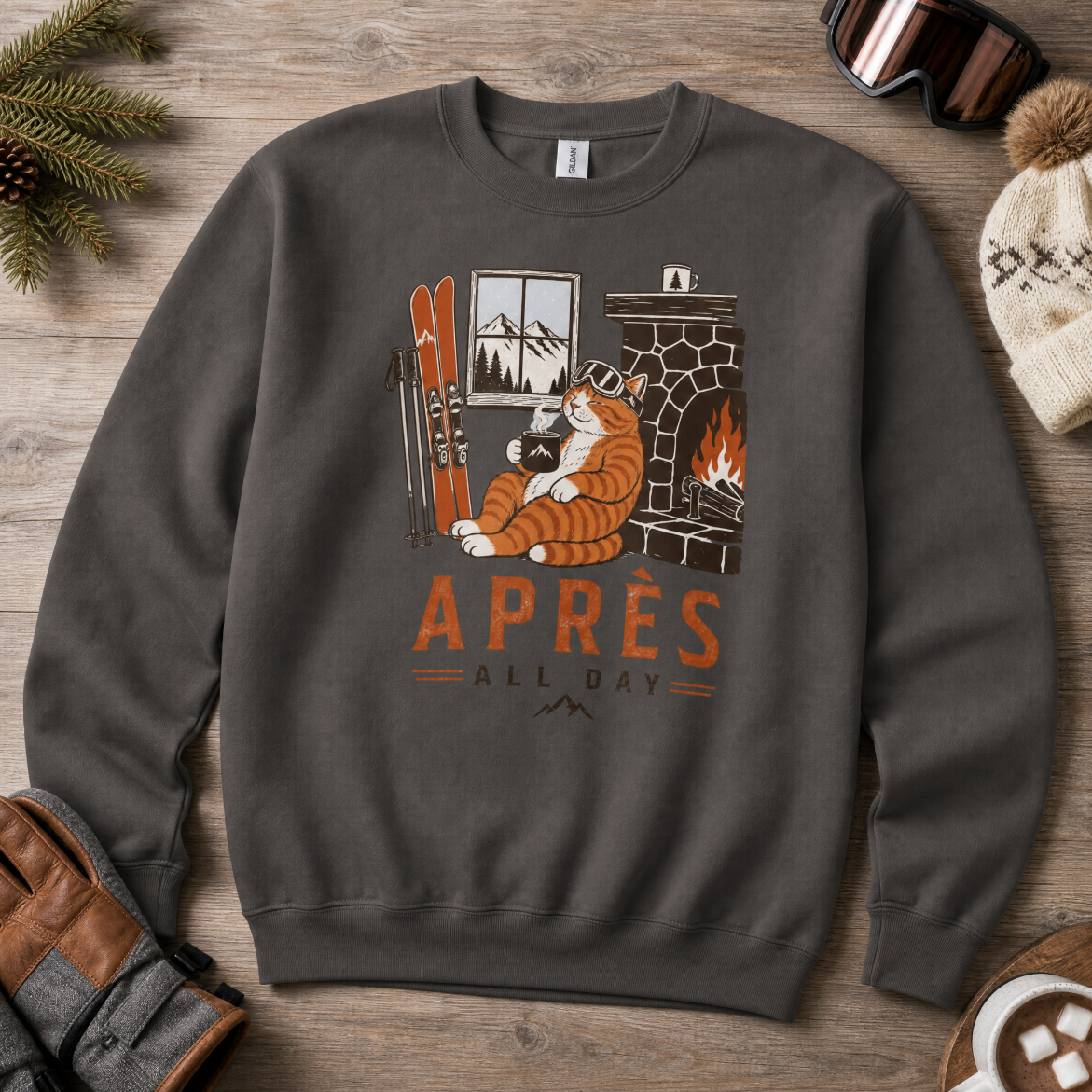 Charcoal gray sweatshirt with a graphic of a cat by a fireplace and 'APRES ALL DAY' text, surrounded by winter accessories on a wooden surface.