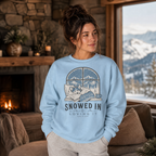 Woman wearing a light blue sweatshirt with a graphic design of a dog and text 'Snowed In & Loving It' in a cozy room with a fireplace and mountain view.