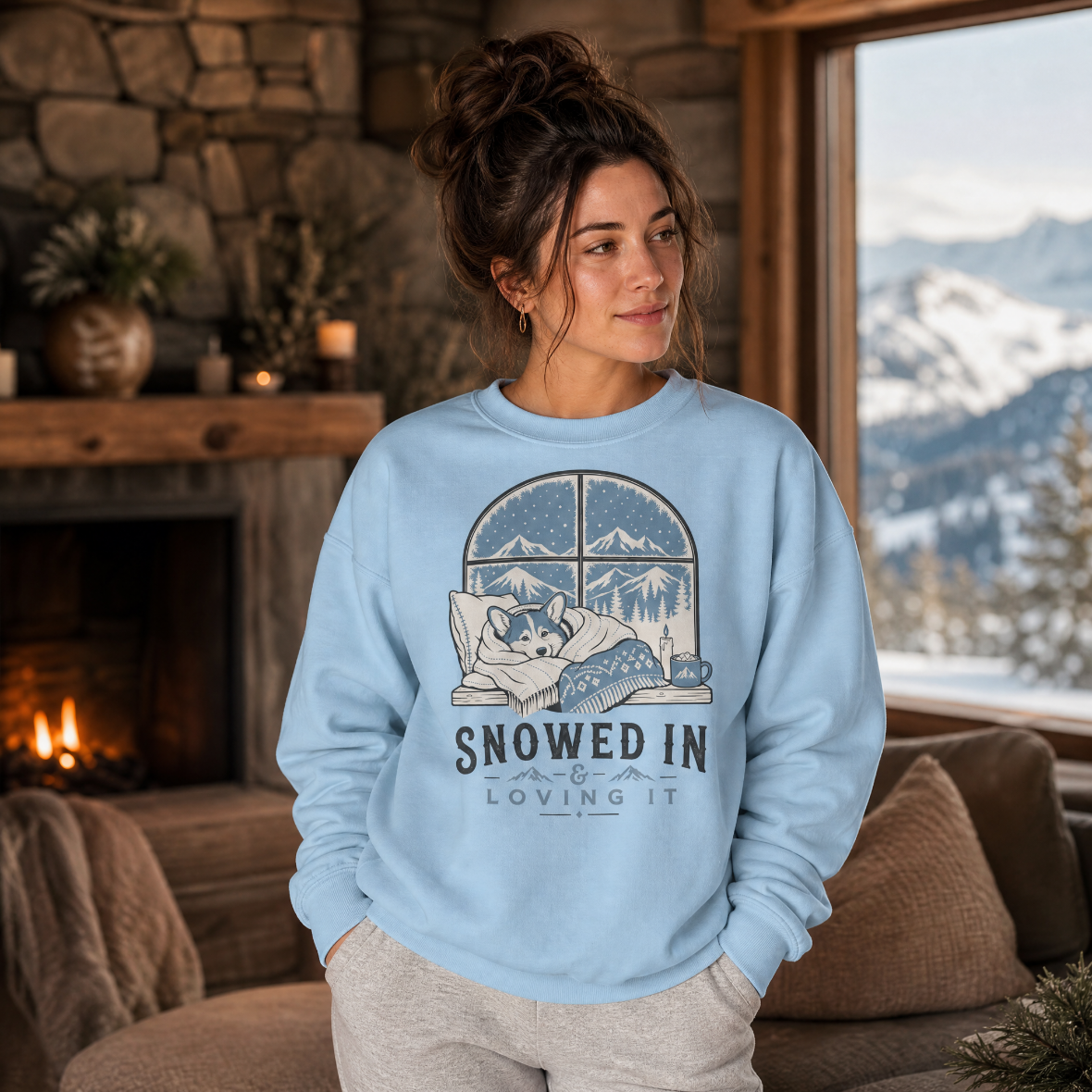 Woman wearing a light blue sweatshirt with a graphic design of a dog and text 'Snowed In & Loving It' in a cozy room with a fireplace and mountain view.