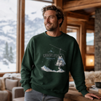 Man wearing a green sweatshirt with a chairlift graphic in a cozy indoor setting with snowy mountains outside.