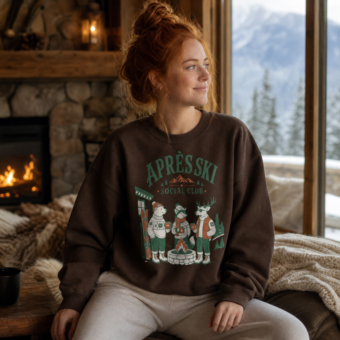 Woman wearing an 'Apres Ski Social Club' sweatshirt sitting by a fireplace with a snowy mountain view.