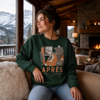 Woman wearing a forest green sweatshirt with a tiger graphic and 'APRES ALL DAY' text, sitting in a cozy room with a fireplace and mountain view.