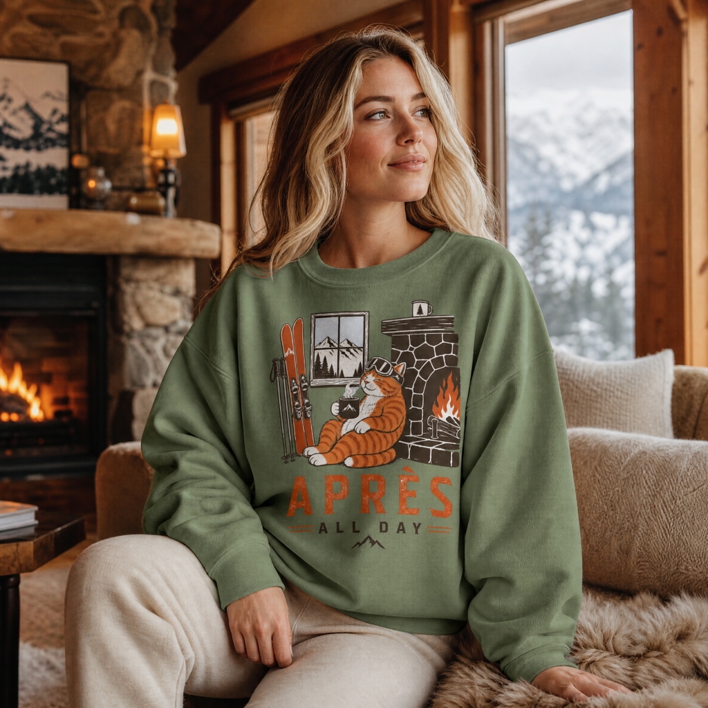 Woman wearing a military green sweatshirt with a graphic design sitting in a cozy room with a fireplace and mountain view.